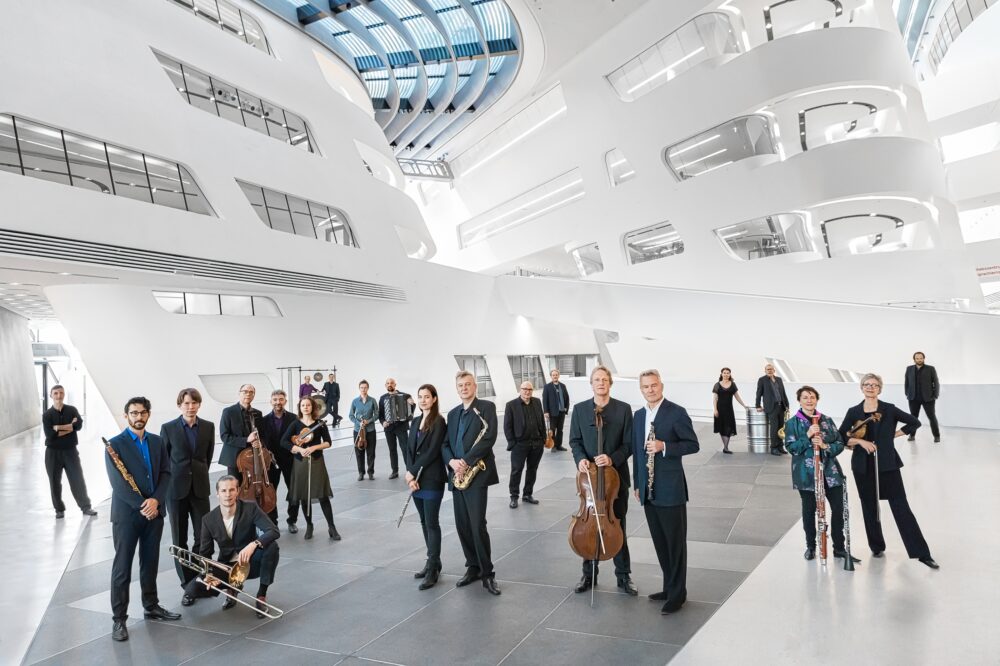 Zu sehen sind die Musiker*innen vom Klangforum Wien. Sie stehen verteilt in dem Foyer eines hellen, futuristischen Gebäudes. Alle Musiker*innen halten ihr Instrument in den Händen.